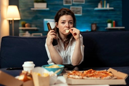 Woman eating snacks alone on a couch, representing unhealthy eating patterns and emotional distress.