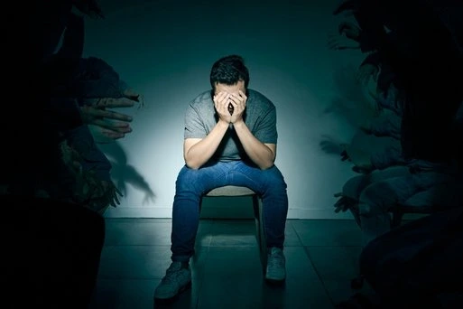 Man sitting alone with head in hands, surrounded by shadowy figures, looking distressed.